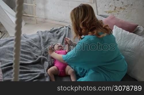Young mother massaging infant baby, doing gymnastic, physical training, exercises for babies in bed. Shot over shoulder. Caring mom developing her baby girl, making gymnastics exercises. Slow motion. Steadicam stabilized shot.