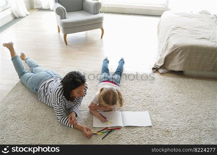 Young mother or babysitter draws with little daughter, lying on warm floor with carpet at home. Mom drawing in sketchbook with small elementary girl on weekend. Art hobby, children’s education.. Mother drawing with little girl daughter, lying on floor at home. Art hobby, children’s education