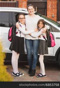 Young mother posing with daughters wearing backpacks next to the car