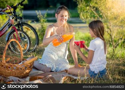 Young mother pouring juice from bottle into daughter&rsquo;s cup on picnic at park