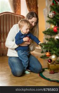 Young mother showing her baby son how to decorate Christmas tree with baubles