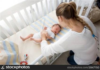 Young mother sitting near the cradle and holding baby&rsquo;s hand