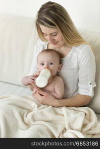 Young mother sitting on bed and feeding her baby from bottle