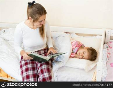 Young mother sitting on bed next to daughter and reading a story