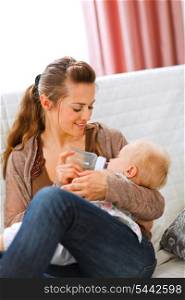 Young mother sitting on sofa and feeding her baby at home&#xA;