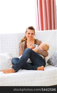Young mother sitting on sofa and feeding her baby &#xA;