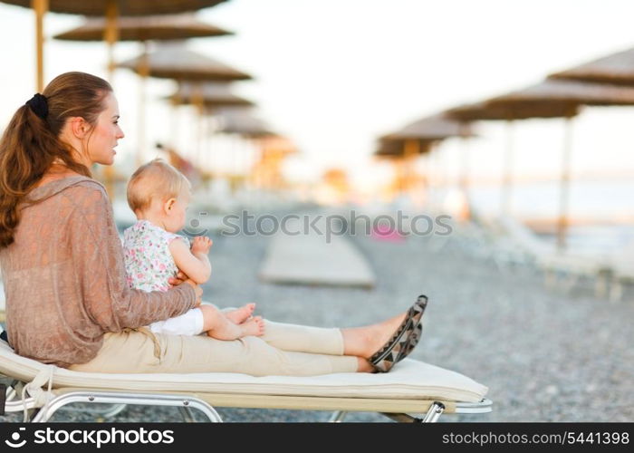 Young mother sitting with baby on sunbed on the beach at evening and looking into the distance