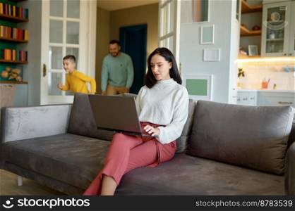 Young mother trying to work on laptop online while father and son playing behind her and making noise. Busy woman working from home in loudly living room sitting on sofa. Young mother trying to work on laptop online while father and son playing