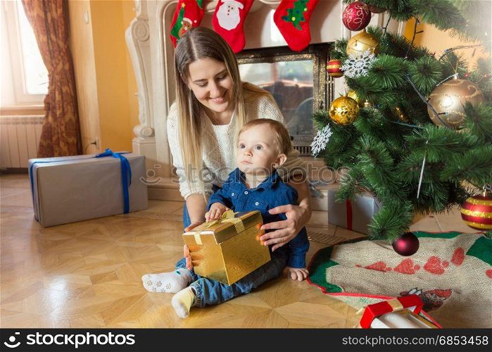 Young mother with her baby son sitting at Christmas tree and looking inside gift box