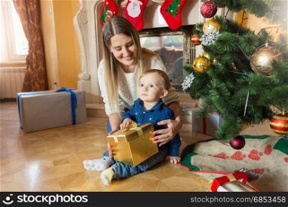 Young mother with her baby son sitting at Christmas tree and looking inside gift box
