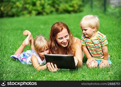 young mother with her children lie on grass and looking at screen of laptop. summer day.