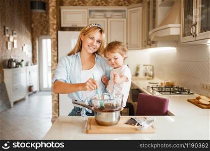 Young mother with her daughter mixing melted chocolate in a bowl. Cute woman and little girl cooking on the kitchen. Happy family prepares sweet dessert at the counter