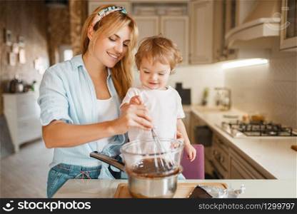 Young mother with her daughter mixing melted chocolate in a bowl. Cute woman and little girl cooking on the kitchen. Happy family prepares sweet dessert at the counter. Mother with her daughter mixing melted chocolate