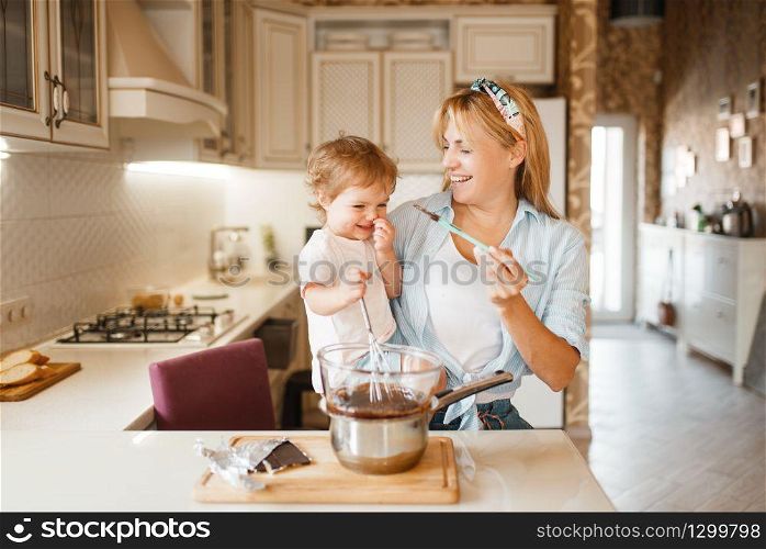 Young mother with her daughter mixing melted chocolate in a bowl. Cute woman and little girl cooking on the kitchen. Happy family prepares sweet dessert. Mother with her daughter mixing melted chocolate