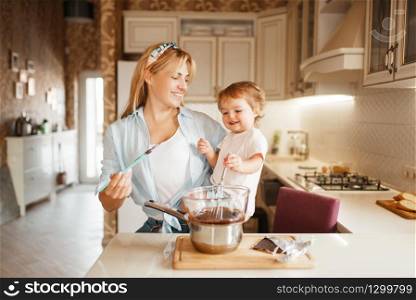 Young mother with her daughter mixing melted chocolate in a bowl. Cute woman and little girl cooking on the kitchen. Happy family prepares sweet dessert. Mother with her daughter mixing melted chocolate