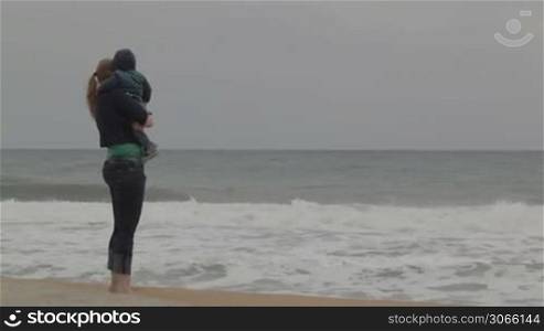 Young mother with her little son staying on the beach and looking at the sea waves. No faces visible.