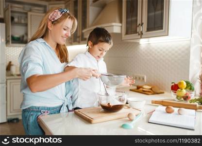 Young mother with her son mixing melted chocolate in a bowl. Cute woman and little boy cooking on the kitchen. Happy family prepares sweet dessert at the counter and having fun