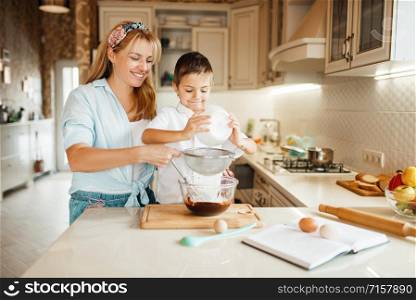 Young mother with her son mixing melted chocolate in a bowl. Cute woman and little boy cooking on the kitchen. Happy family prepares sweet dessert at the counter and having fun. Mother with son mixing melted chocolate in a bowl