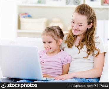 Young mother with little daughter sitting together at home and looking at laptop