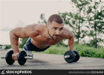 Young motivated sportsman stands in plank pose, trains muscles with barbells, focused into distance with determined expression, has naked torso, has strong muscular body. Athlete bodybuilder outdoor