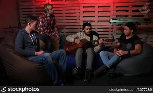 Young musicians jamming in garage. Teenage music band performing music unplugged in garage over musical instruments background. Stylish guitarist playing acoustic guitar, drummer setting the rhythm and the rest of group members singing song.