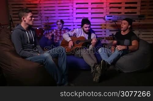Young musicians rehearsing unplugged together in garage. Attractive teenagers sitting comfortably on chill bags on the stage. Stylish guitarist playing acoustic guitar while other band members singing along.