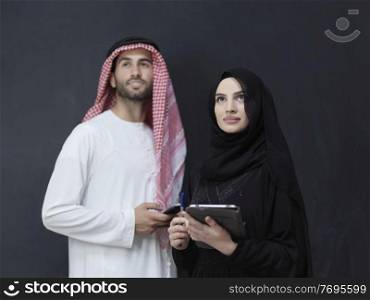 Young muslim business couple using technology devices. Successful Arabs in traditional clothes standing in front of black chalkboard.