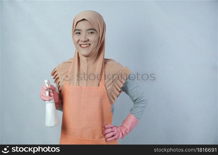 Young muslim housewife with pink rubber gloves in apron holding cleaning spray bottle on grey background