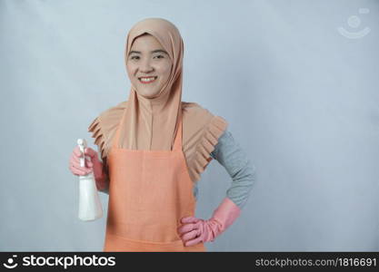 Young muslim housewife with pink rubber gloves in apron holding cleaning spray bottle on grey background