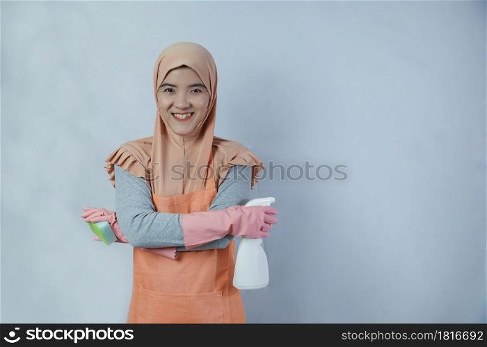 Young muslim housewife with pink rubber gloves in apron holding cleaning spray bottle and sponge on grey background