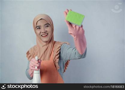 Young muslim woman housewife with pink rubber gloves in apron holding cleaning spray bottle and sponge on grey background