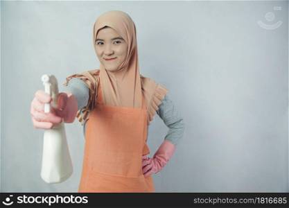 Young muslim woman housewife with pink rubber gloves in apron holding cleaning spray bottle on grey background