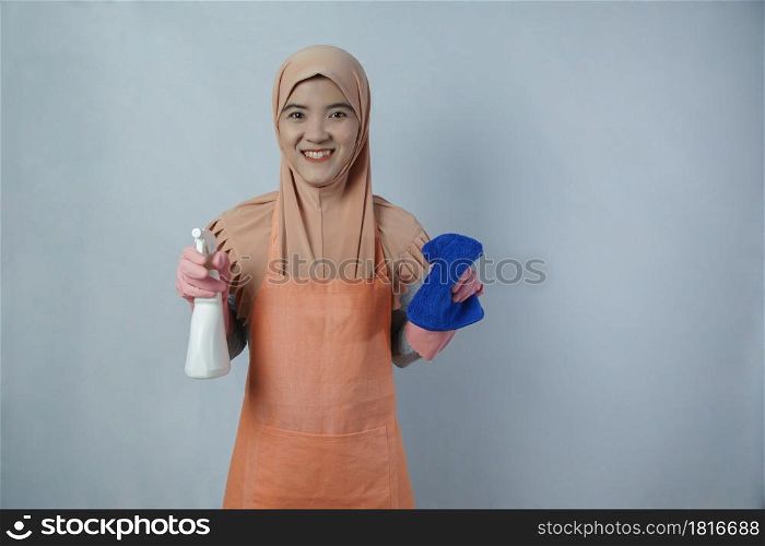 Young muslim woman housewife with pink rubber gloves in apron holding cleaning spray bottle and rag on grey background