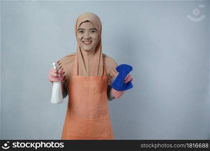 Young muslim woman housewife with pink rubber gloves in apron holding cleaning spray bottle and rag on grey background