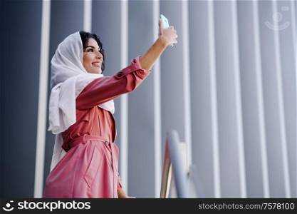 Young Muslim Woman wearing hijab headscarf photograph with a smartphone in urban background. Muslim Woman with hijab taking selfie with smartphone