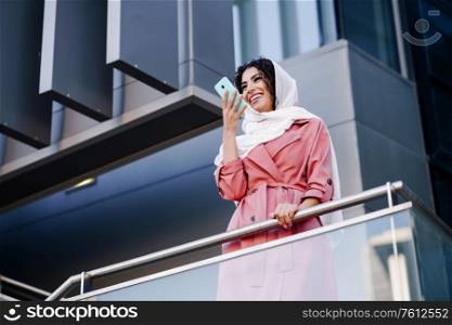 Young Muslim Woman wearing hijab headscarf recording voice note with a smartphone in a office building. Business background.. Muslim Woman with hijab recording voice note with a smartphone.