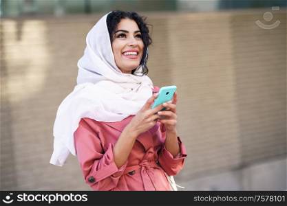 Young Muslim woman wearing hijab headscarf texting message with her smartphone in urban background.. Young Muslim woman wearing hijab texting message with her smartphone.