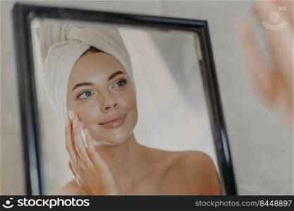 Young naked European woman touches soft skin on face, looks at herself in bathroom mirror, wears wrapped white towel on head after taking bath, poses bare shoulders. Beauty and skin care concept