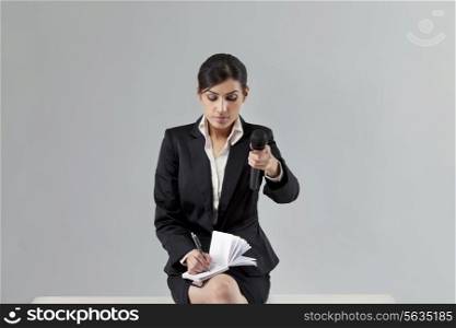 Young news reporter holding out microphone while writing in notepad