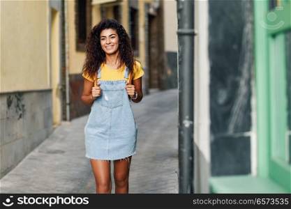 Young North African tourist woman with black curly hairstyle outdoors. Arab traveler girl in casual clothes in the street. Happy female wearing yellow t-shirt and denim dress in urban background.