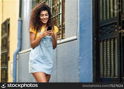 Young North African woman walking on the street looking at her smart phone. Smiling Arab girl in casual clothes with black curly hairstyle.. North African woman walking on the street looking at her smart phone