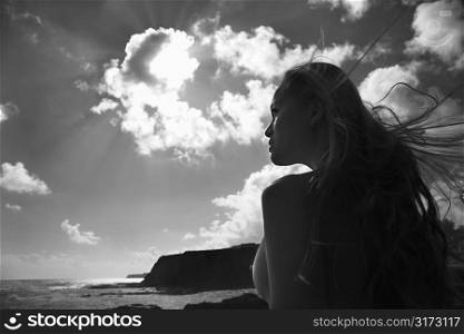 Young nude Asian woman looking out towards ocean with hair blowing in wind.