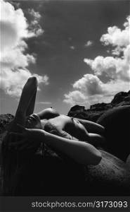 Young nude Asian woman lying on back on rocks with hands to head.