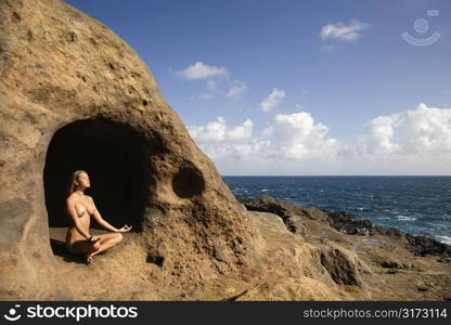 Young nude Asian woman mediatating in cave with coastline of Maui, Hawaii.