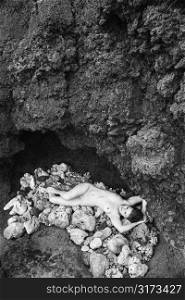 Young nude Caucasian woman lying down on pile of rocks with hands above head.