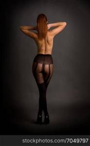 young nude girl in pantyhose and shoes is standing with her back on a black background