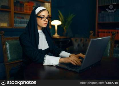 Young nun in a cassock and glasses works on laptop. The sister in the monastery, religion and faith, religious people. Young nun in a cassock and glasses works on laptop