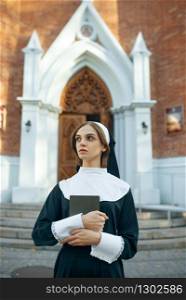Young nun in a cassock holds a book, church on background. The sister is preparing for prayer in the monastery, religion and faith, religious person. Young nun in a cassock, church on background
