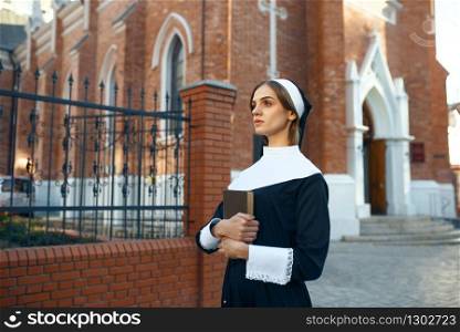 Young nun in a cassock holds a book, church on background. The sister is preparing for prayer in the monastery, religion and faith, religious people. Young nun in a cassock, church on background