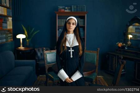 Young nun in a cassock with a cross around her neck holds a book. The sister is preparing for prayer in the monastery. Young nun in a cassock holds book